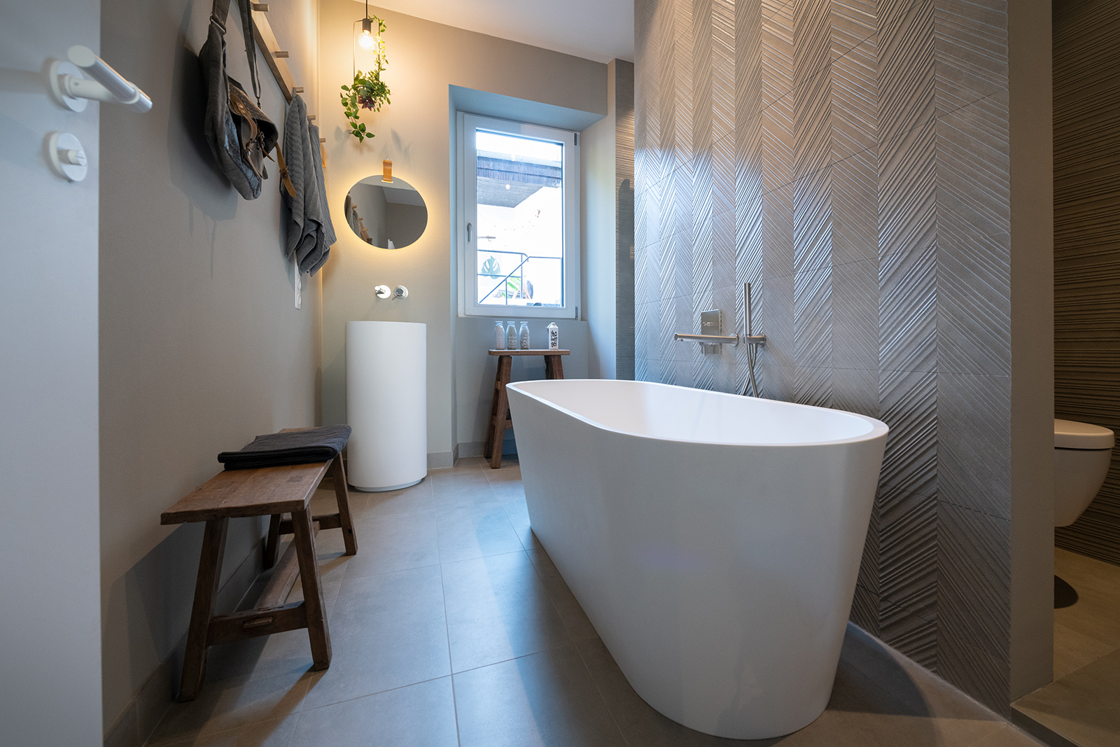 Übersicht Bad: Freistehende Badewanne vor Strukturfliesen und stimmunngsvoll beleuchteter Waschbereich